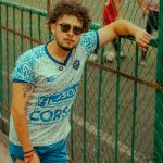 Stylish man in sportswear with tattoo leaning on a fence by a soccer field.